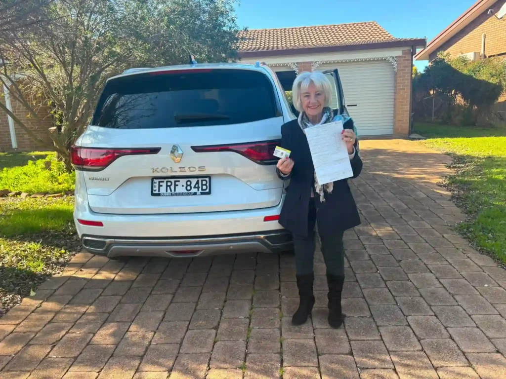 A senior female driver smiling and holding her passed driver assessment certificate next to her car in Dubbo NSW with AUZ Driving School